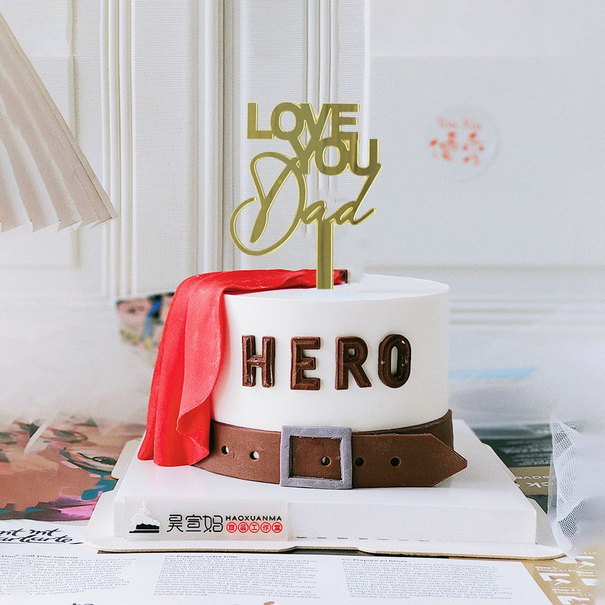A white cake with a round shape features a decorative cake topper that reads "LOVE YOU Dad" in gold lettering. The cake is adorned with a red cape draped over one side, and the word "HERO" is spelled out in brown letters around the middle. The cake is placed on a white cake board, accompanied by a design that suggests a festive or celebratory atmosphere. The background includes light-colored elements, enhancing the overall presentation.