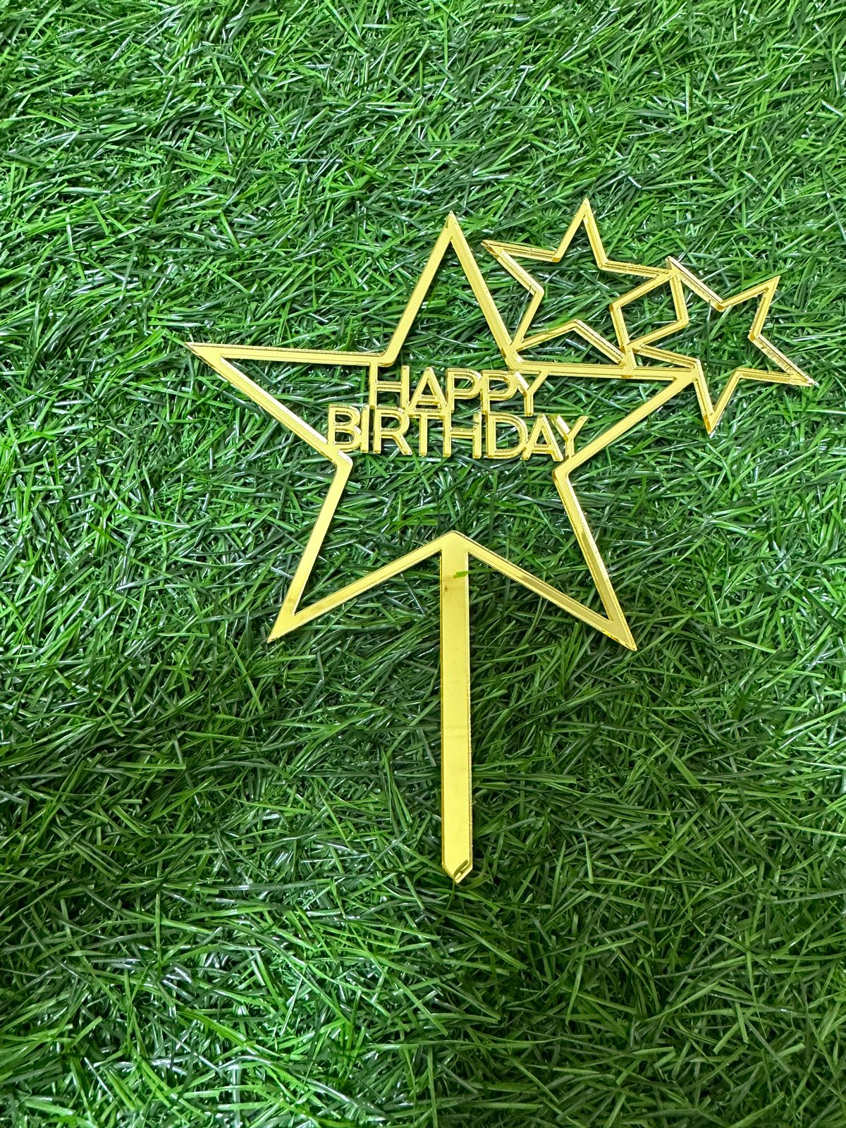 The image shows a large gold star-shaped cake topper with the words "HAPPY BIRTHDAY" prominently displayed in the center. Below the main star, there is a stick for inserting it into a cake. Additionally, there are two smaller gold star outlines positioned next to the main star. The backdrop features a green artificial grass surface.