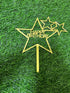 The image shows a large gold star-shaped cake topper with the words "HAPPY BIRTHDAY" prominently displayed in the center. Below the main star, there is a stick for inserting it into a cake. Additionally, there are two smaller gold star outlines positioned next to the main star. The backdrop features a green artificial grass surface.
