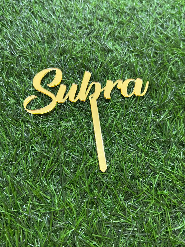 An elegant gold cake topper with the name "Subra" written in a cursive script. The topper has a thin stake for insertion into cakes. It is placed against a background of green artificial grass, providing a vibrant contrast to the gold color.