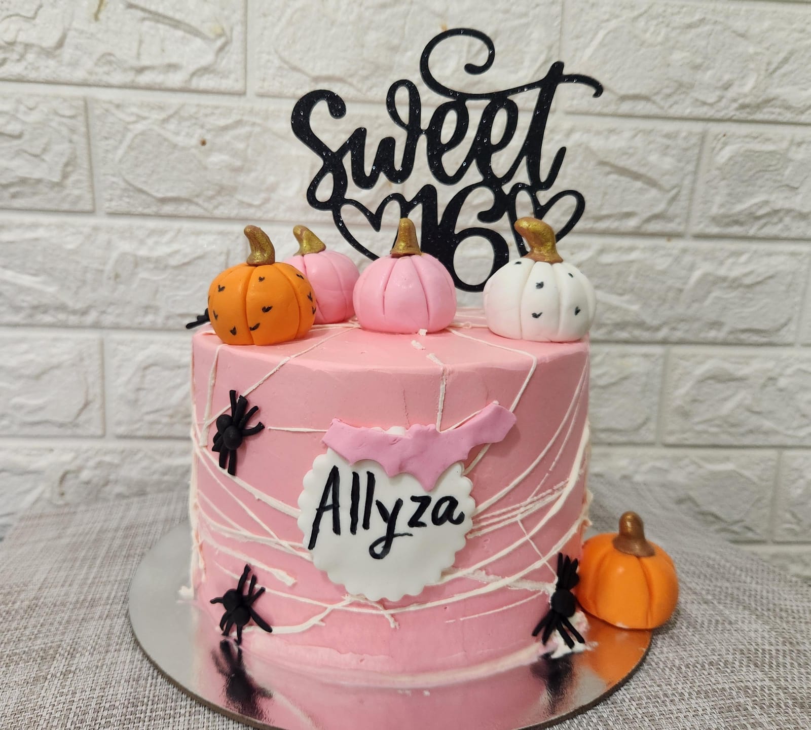A tiered pink birthday cake adorned with a black cardstock topper that says "Sweet 16." The cake is decorated with small fondant pumpkins in orange and white, and has black spider decorations on the sides. A pink flag with the name "Allyza" is attached to the front of the cake. The background features a light-colored, textured wall.