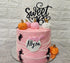 A tiered pink birthday cake adorned with a black cardstock topper that says "Sweet 16." The cake is decorated with small fondant pumpkins in orange and white, and has black spider decorations on the sides. A pink flag with the name "Allyza" is attached to the front of the cake. The background features a light-colored, textured wall.