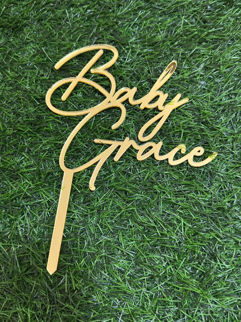 The image features a customized acrylic cake topper designed for celebrations. The topper is shaped in an elegant script font that reads "Baby Grace." It has a shiny gold finish and includes a tapered spike at the bottom for easy insertion into a cake. The background consists of green grass or faux grass, enhancing the visual presentation of the cake topper.