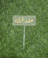 A clear acrylic cake topper with a rectangular shape, featuring elegant gold lettering that reads in Arabic. It is mounted on a thin stick for easy placement on a cake. The topper is set against a backdrop of artificial green grass.