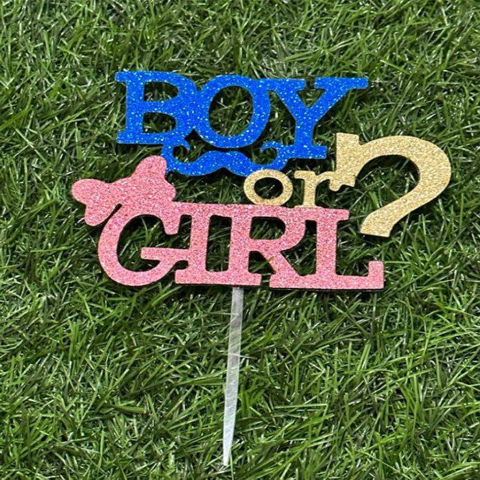 A customized cardstock cake topper reading "BOY or GIRL?" with colorful glitter. The word "BOY" is in blue with a blue mustache graphic, while "GIRL" is in pink, accompanied by a pink bow. The text is mounted on a clear stick, set against a green grass background.