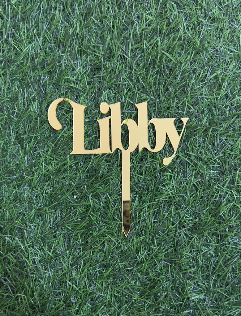 A gold acrylic cake topper featuring the name "Libby" in an elegant cursive font. The topper is placed on a background of green grass, highlighting its shiny surface and design. The topper has a pointed base for easy insertion into a cake.