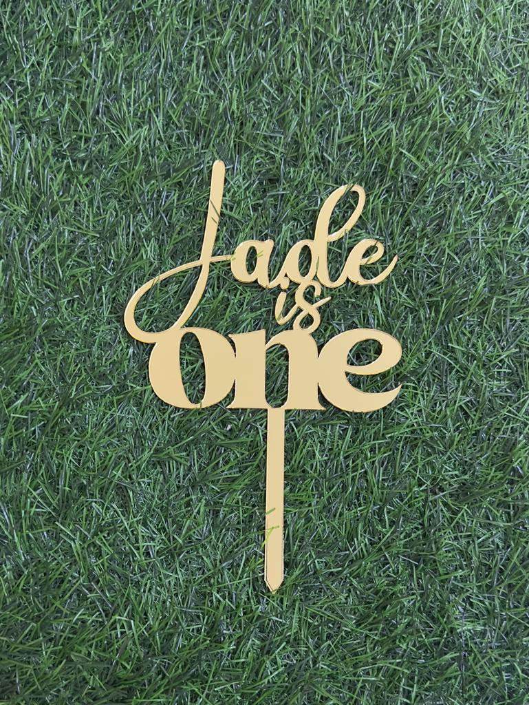 A gold acrylic cake topper is displayed on a green grass-like surface. The topper features the text "Jade is One" in an elegant, flowing script, with "Jade" in a larger font size than the rest of the text. The design includes a stake for inserting into a cake.