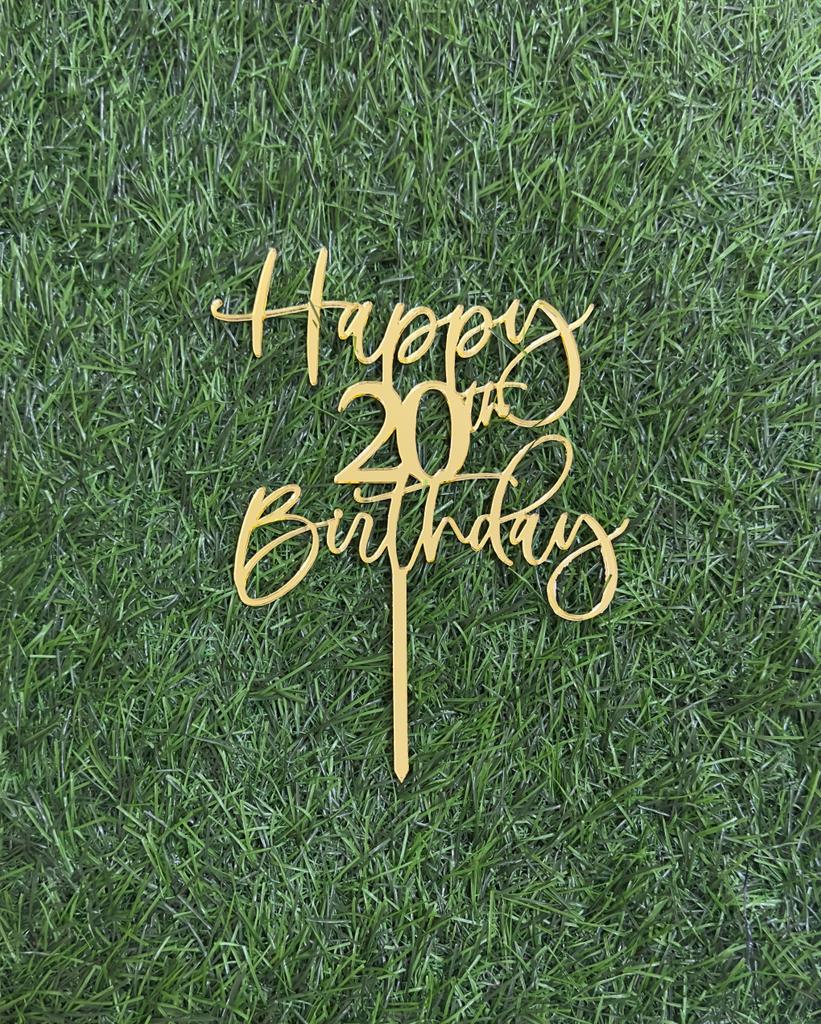 A golden acrylic cake topper featuring the phrase "Happy 20th Birthday" in a decorative, cursive font. The topper is placed on a background that resembles green grass, highlighting its design.