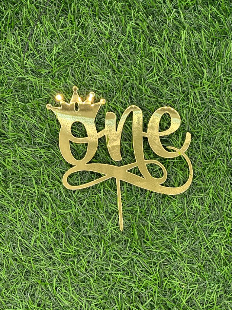 The image features a gold acrylic cake topper that reads "one" in a stylized, cursive font. Above the text, there is a small crown design, enhancing the celebratory theme. The topper is placed against a background of green grass, adding a vibrant contrast.