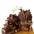A chocolate cake with a glossy drip effect, topped with various chocolate pieces, round candies, and a decorative acrylic cake topper that reads "SIXTY." The cake is positioned next to a stack of chocolate fudge pieces. The background is a golden surface, enhancing the festive appearance of the dessert.