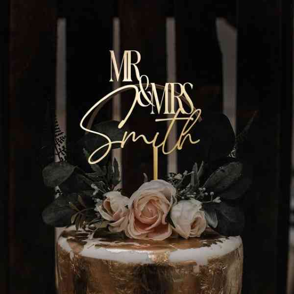 A cake topper featuring the phrase "MR & MRS" in an elegant font, along with the name "Smith." The topper is placed atop a tiered cake adorned with soft pink roses and greenery, including dark leaves. The cake has a gold finish, adding a touch of sophistication to the overall presentation. The background is dark, enhancing the visual impact of the cake and topper.