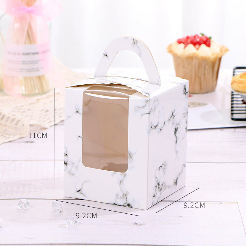 A white cupcake box with a marble design and a handle, featuring a transparent window on the front. The box dimensions are labeled: 11 cm in height and 9.2 cm in width and depth. It is placed on a light wooden surface with decorative items in the background, including a cupcake topped with strawberries and a glass jar with a pink ribbon. Small decorative crystals are scattered around the box.