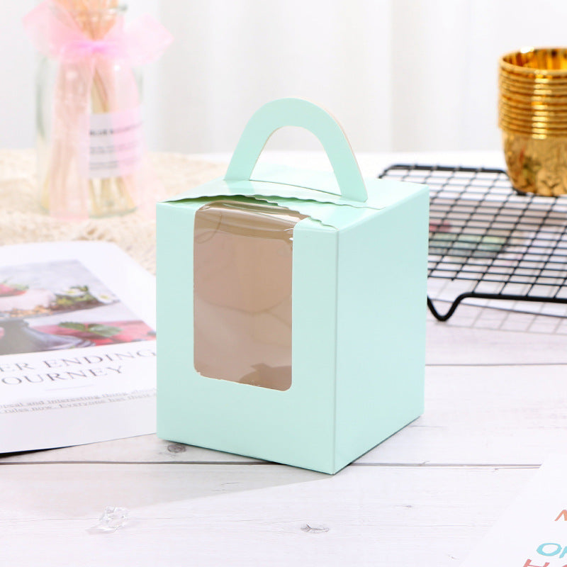 A mint green cupcake box designed for single portions is displayed, featuring a clear plastic window on the front. The box is topped with a handle and has a scalloped edge. In the background, there are blurred decorative elements, including a pink ribbon and a gold container, along with a magazine or paper laid flat on a wooden surface.