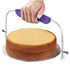 A cake leveler is shown in the image, featuring a person?s hand gripping the handle. The tool has a curved metal frame with two adjustable wires on either side. It is positioned above a round, beige cake placed on a silver cake board. The cake has a flat top and slightly rounded edges, indicating it is ready to be leveled. The handle is purple in color, providing a comfortable grip for use.