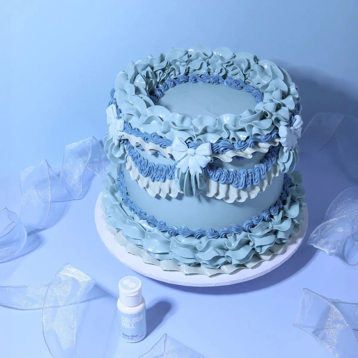 Elegant blue cake with intricate ruffled icing and bows on a light blue background. Includes a small "baby blue" food coloring bottle, creating a serene mood.