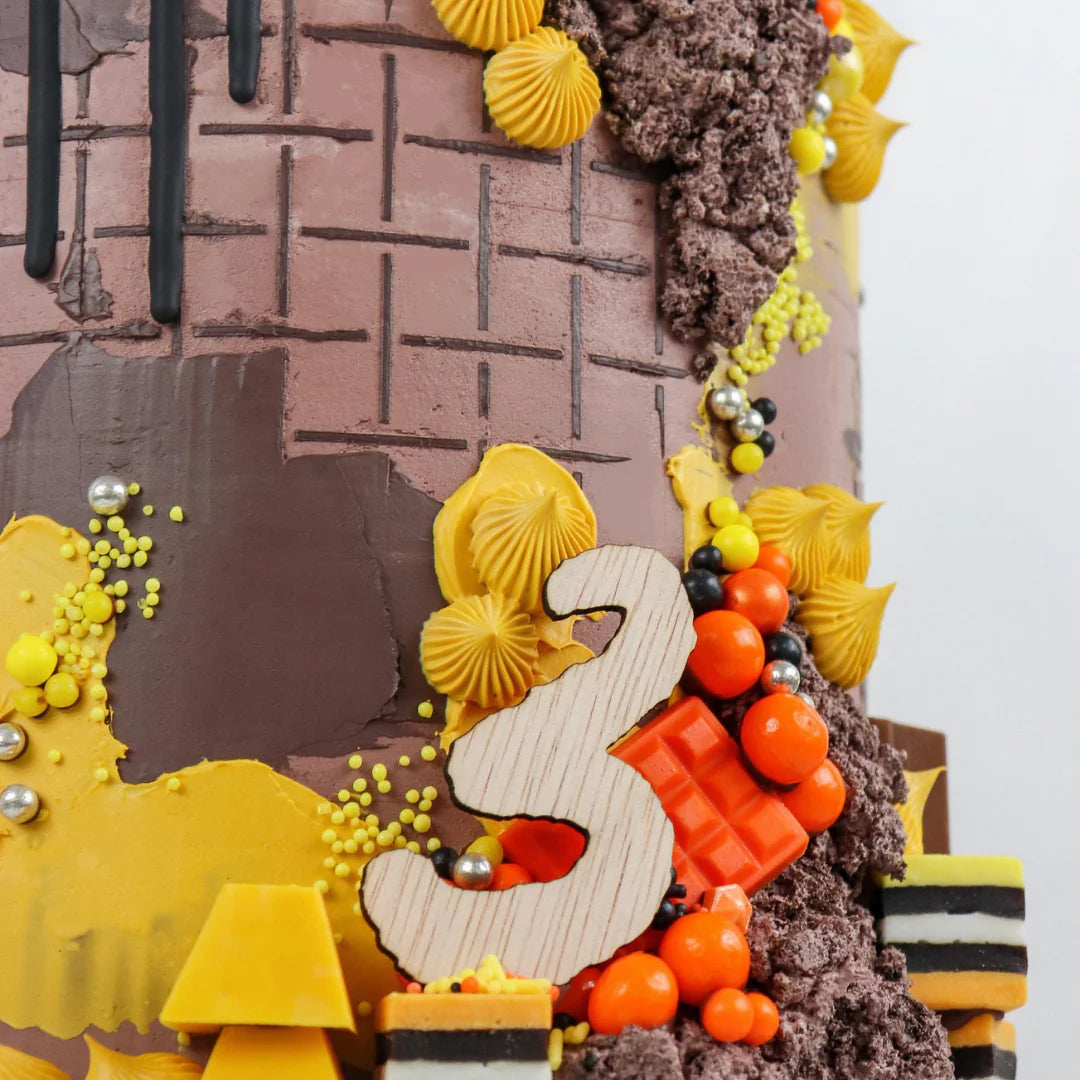 Close-up of a decorative cake featuring a textured brown and yellow design, a wooden number 3, and colorful candy accents, creating a festive atmosphere.