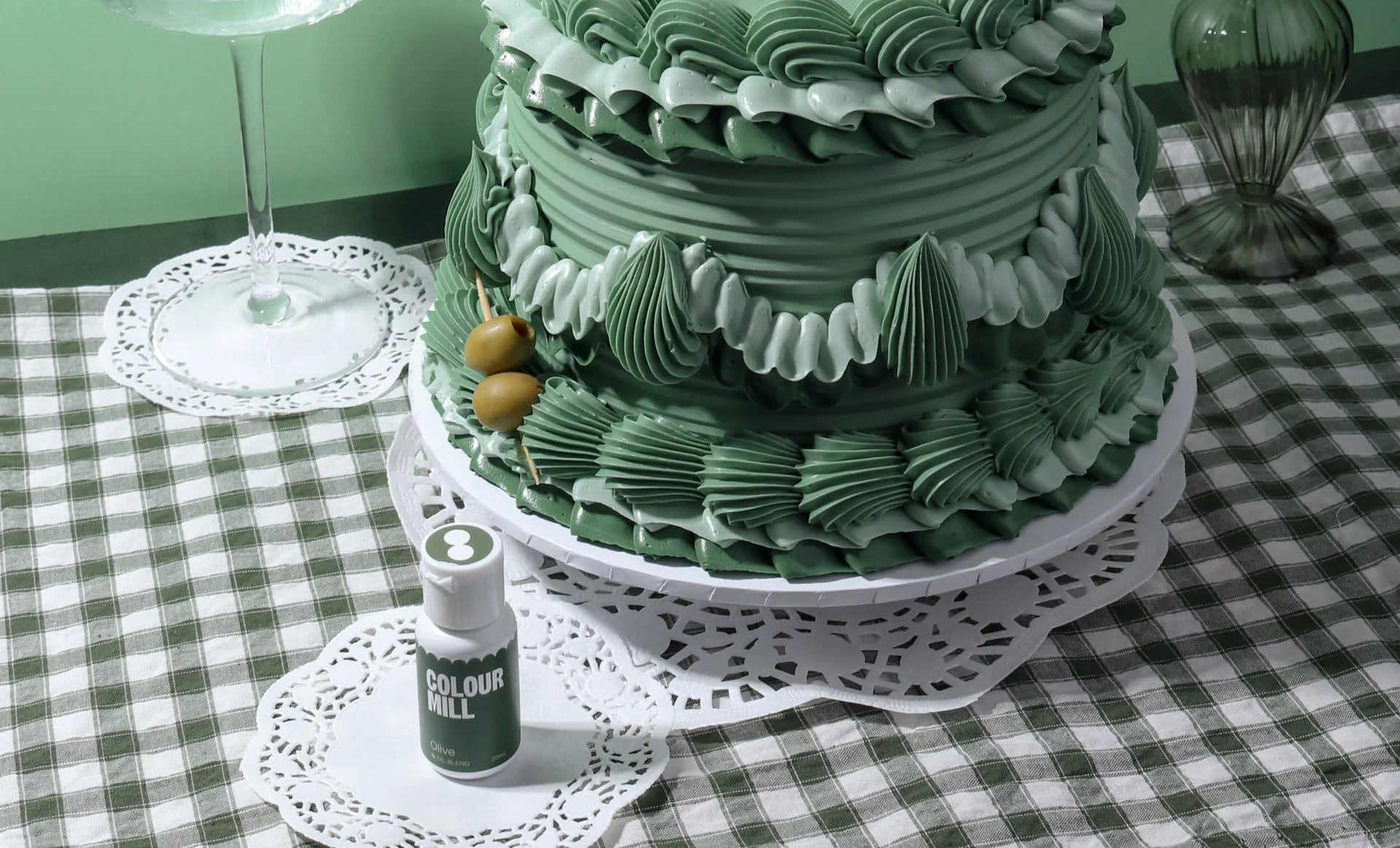 Elegant green cake with intricate frosting patterns on a white stand, set on a green checkered tablecloth. A small bottle labeled "Colour Mill" sits nearby.