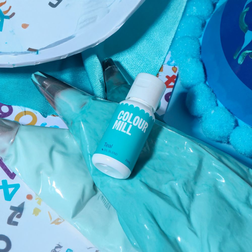 Bottle of teal color mill dye on a teal background with matching frosting bags, a paper plate with colorful numbers, and blue pom-poms, conveying a crafty, vibrant tone.