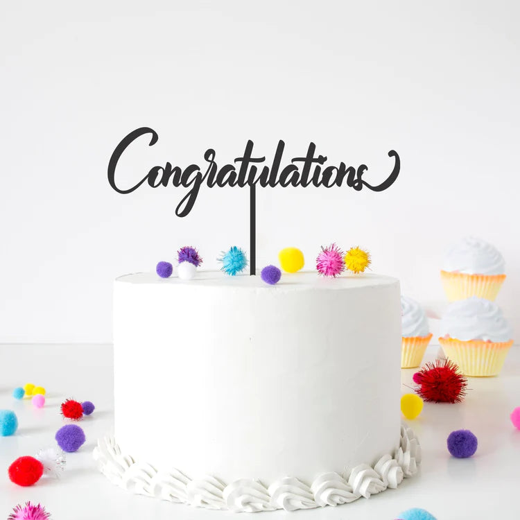 A white frosted cake adorned with a black acrylic topper that reads "Congratulations." The cake is decorated with colorful pom-poms in various colors scattered on top and around the base. In the background, there are small cupcakes with light blue frosting and yellow cupcake liners.
