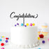 A white frosted cake adorned with a black acrylic topper that reads "Congratulations." The cake is decorated with colorful pom-poms in various colors scattered on top and around the base. In the background, there are small cupcakes with light blue frosting and yellow cupcake liners.