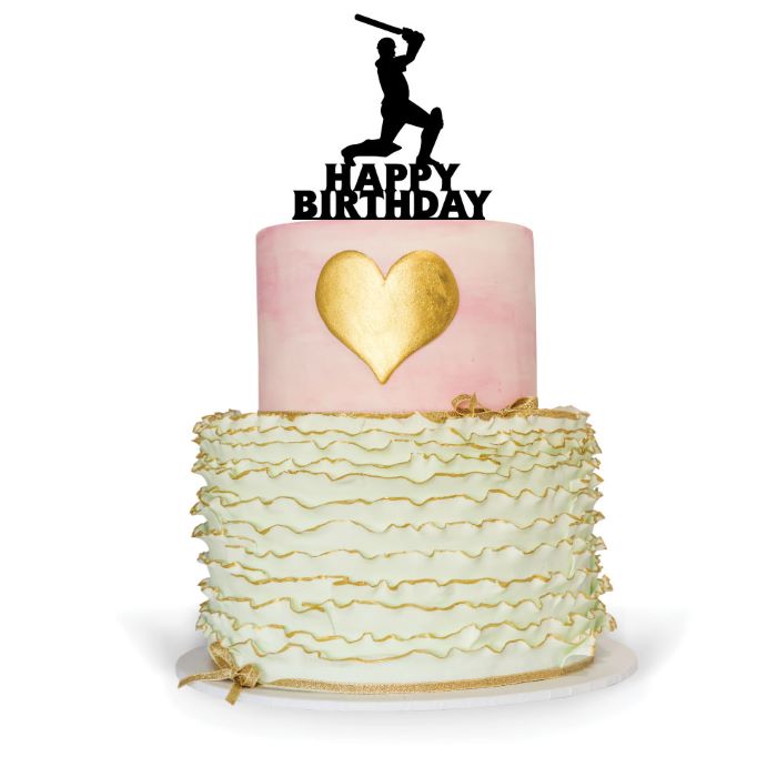 A two-tier birthday cake featuring a pink top layer with a golden heart design and a white, ruffled bottom layer. On top of the cake is a black silhouette of a cricketer holding a bat, along with the text "HAPPY BIRTHDAY" in bold letters. The cake is decorated with a gold ribbon around the base.