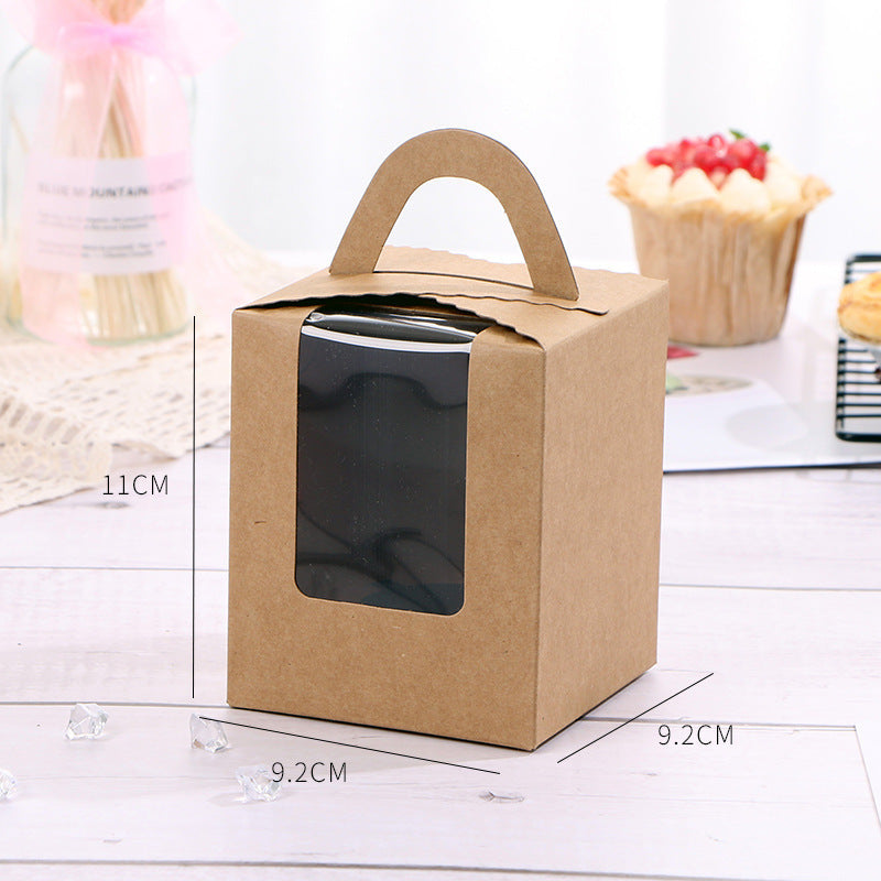The image features a single portion cupcake box made of brown kraft paper. The box has a rectangular shape with a handle on top for easy carrying. It includes a transparent plastic window on one side, allowing a view of the contents inside. The dimensions of the box are indicated, measuring 11 cm in height and 9.2 cm in width on each side. In the background, there are some decorative items, including a cupcake with frosting and berries, and some small crystal-like decorative pieces scattered on the surface.