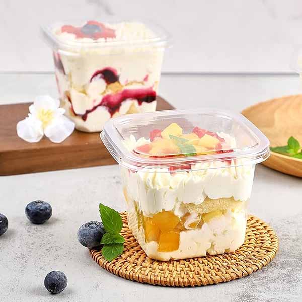 Two square plastic dessert cups, each with a clear lid, are filled with layered desserts. The front cup features a mixture of cream, fruit pieces, and cake, topped with whipped cream and fruit. A mint leaf decorates the top. The back cup displays a dessert with a darker sauce and more layers. Blueberries scatter around the cups, and a white flower lies nearby, set on a light grey surface with a wooden plate in the background. The image conveys a fresh and inviting presentation of sweet treats.