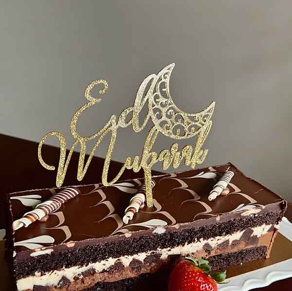 A chocolate cake with a decorative topping that reads "Eid Mubarak" in a glittery gold font. The topper features a crescent moon design. The cake has a layered appearance with a dark chocolate base, a light cream layer, and is garnished with swirled chocolate decorations and two striped candies. A fresh strawberry sits on the side of the cake.