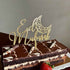 A chocolate cake with a decorative topping that reads "Eid Mubarak" in a glittery gold font. The topper features a crescent moon design. The cake has a layered appearance with a dark chocolate base, a light cream layer, and is garnished with swirled chocolate decorations and two striped candies. A fresh strawberry sits on the side of the cake.