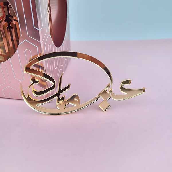 A golden acrylic cake topper featuring the text "??? ?????" (Eid Mubarak) in an elegant, stylized font. The topper is positioned in front of a pink cake box with a geometric design, against a light blue background. The scene emphasizes the festive theme associated with Eid celebrations.