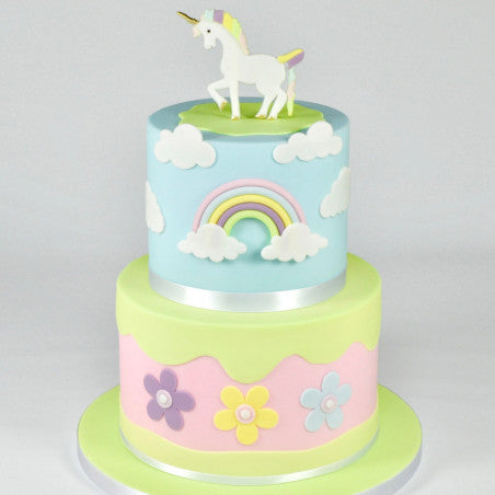 A two-tiered cake decorated in a vibrant, whimsical style. The top tier features a light blue surface with a rainbow and fluffy white clouds, topped with a fondant unicorn figurine. The bottom tier is light green with a pink wavy band embellished with colorful flower decorations. The overall theme is playful and cheerful, ideal for a celebration.