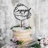 A simple layered cake displayed on a glass stand with a textured, white icing on top. Atop the cake, there is a black acrylic cake topper featuring the words "Engaged & Frank Kate" surrounded by decorative leaf designs, and the date "09.30.2022" below. The cake is adorned with green leaves and small purple flowers. In the background, there are two tall, cream-colored candles and a softly blurred backdrop.