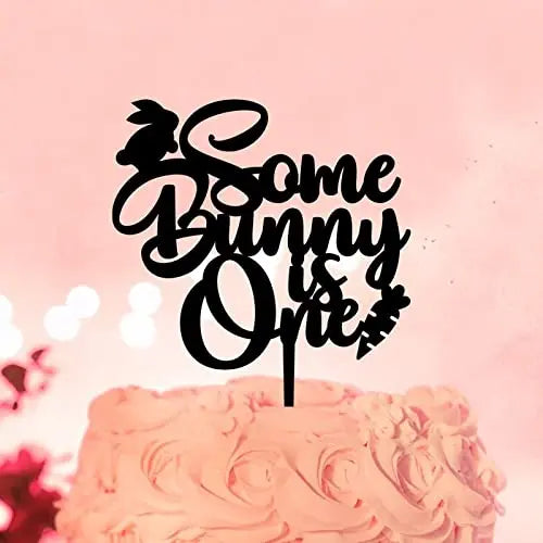 The image features a black cake topper designed for a first birthday celebration. The topper includes the text "Some Bunny is One," with decorative elements like a bunny silhouette and a carrot. It is placed on a pink-frosted cake, with a soft, blurred pink background adding a festive touch.