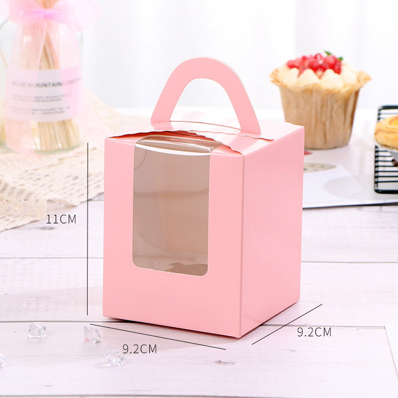 A pink cupcake box with dimensions 11 cm in height and 9.2 cm in width and depth. The box features a handle on top and a clear plastic window on the front, allowing visibility of the cupcake inside. In the background, there are decorative elements including a jar with a pink ribbon and a couple of baked goods, such as a cupcake topped with fruit and a tart displayed on a wire rack. Small decorative crystals are scattered on the surface.