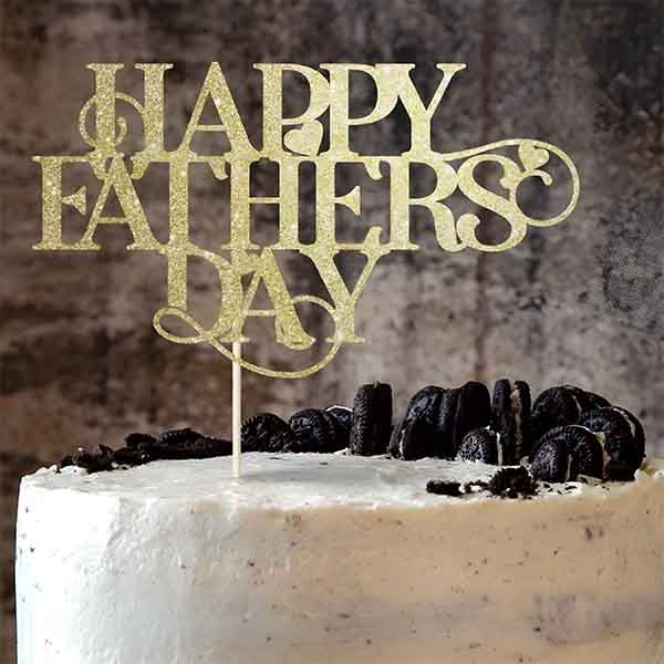A cake with a smooth white frosting is topped with a decorative cake topper that reads "HAPPY FATHERS DAY" in an elegant golden script. The cake is adorned with black cookie crumbs and whole black Oreo cookies around the edges. The background features a textured, dark surface that contrasts with the cake and topper.