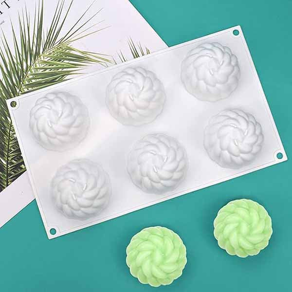 A white silicone mold shaped like chrysanthemum flowers, featuring six cavities, rests on a teal surface. Two finished flower-shaped items, one green and one light green, are placed nearby. A tropical leaf design is visible in the background.