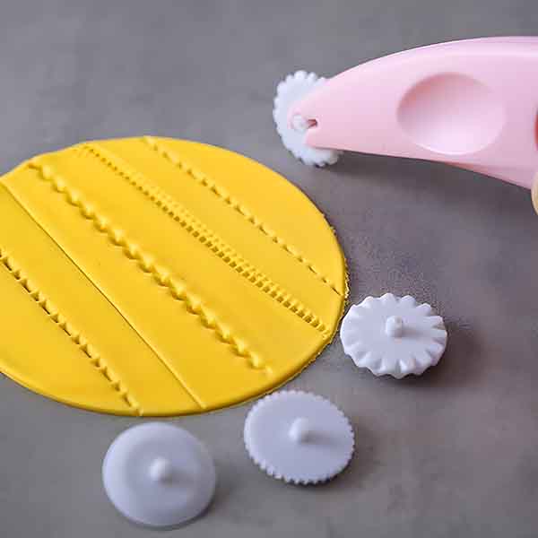 A yellow fondant circle with embossed patterns lies on a gray surface. A pink fondant cutter embosser is in use, featuring a white circular wheel with a serrated edge. Two additional white cutter wheels with different designs rest next to the fondant.