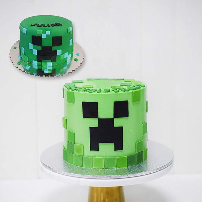 The image features a two-tiered cake designed to resemble a character from a popular video game. The cake is predominantly green and features a pixelated face with black squares representing eyes and a mouth. The top of the cake is decorated with smaller green and blue squares, adding to the pixelated design. It sits on a round silver cake board and is placed on a golden cake stand, against a plain white background.