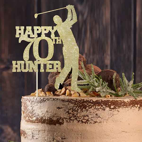A cake topped with a personalised glitter cardstock cake topper featuring the text "HAPPY 70th HUNTER" alongside a silhouette of a golfer in mid-swing. The cake has a rustic, layered appearance and is decorated with nuts, chocolate pieces, and greenery. The background consists of wooden textures.
