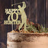 A cake topped with a personalised glitter cardstock cake topper featuring the text "HAPPY 70th HUNTER" alongside a silhouette of a golfer in mid-swing. The cake has a rustic, layered appearance and is decorated with nuts, chocolate pieces, and greenery. The background consists of wooden textures.