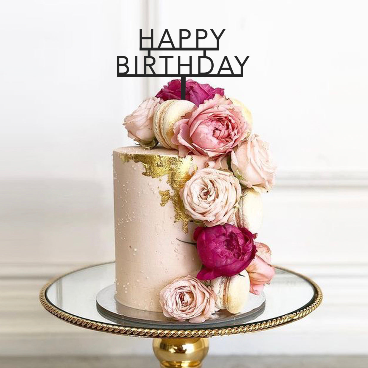 A beautifully decorated birthday cake is centered on a round gold serving platter. The cake is smooth and pastel pink, adorned with delicate flowers including pale pink roses and deep pink peonies, as well as macarons. Atop the cake is a decorative cake topper that reads "HAPPY BIRTHDAY" in bold, black letters. The background is softly blurred, enhancing the cake&