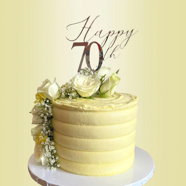 A yellow frosted cake featuring a smooth, layered design. Atop the cake, there is an acrylic topper that reads "Happy 70th" in an elegant script. The cake is adorned with fresh white roses and small green leaves, along with sprigs of baby?s breath flowers, creating a festive and celebratory appearance. The backdrop is a soft, light color, enhancing the cake&