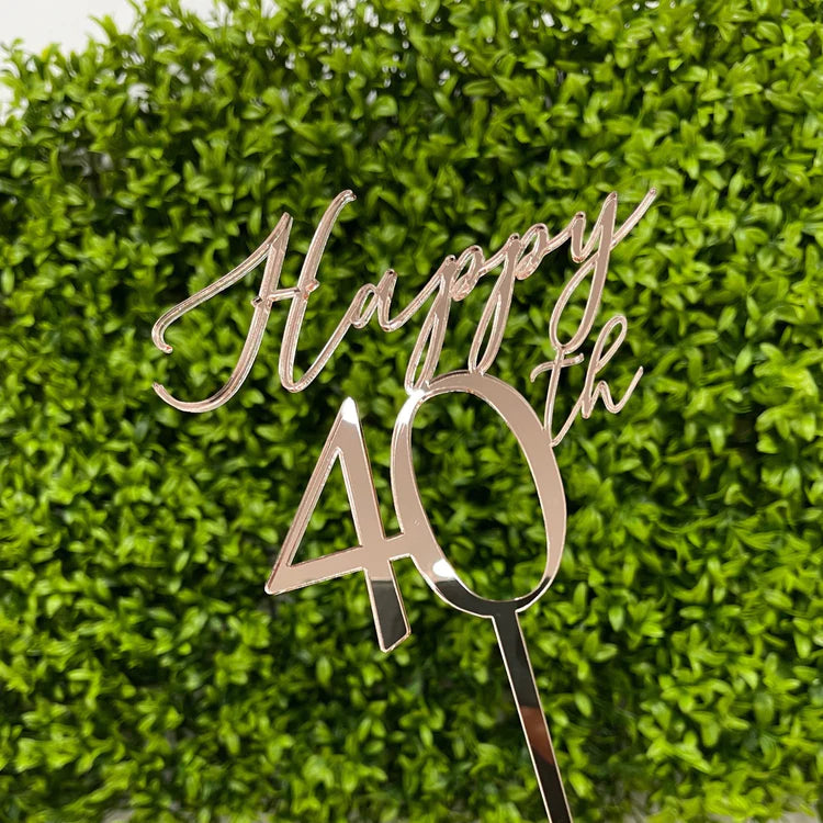 An acrylic cake topper featuring the text "Happy 40th" in a decorative script font. The topper is designed with a shiny rose gold finish and includes a pointed base for easy insertion into a cake. The background consists of green leaves, enhancing the visual appeal of the product.