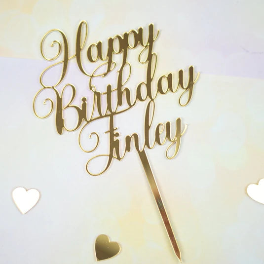 The image features a gold acrylic cake topper with the inscription "Happy Birthday Finley." The lettering is in a decorative, cursive style, and a pointed base is attached for insertion into a cake. The background is a soft, light-colored surface, and there are small heart-shaped decorations scattered around the topper.