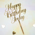 The image features a gold acrylic cake topper with the inscription "Happy Birthday Finley." The lettering is in a decorative, cursive style, and a pointed base is attached for insertion into a cake. The background is a soft, light-colored surface, and there are small heart-shaped decorations scattered around the topper.