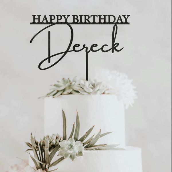A white multi-tier cake decorated with succulents and flowers, topped with a black acrylic cake topper that reads "HAPPY BIRTHDAY" above the name "Dereck." The backdrop is light and neutral, enhancing the decorative elements of the cake and topper.