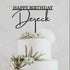A white multi-tier cake decorated with succulents and flowers, topped with a black acrylic cake topper that reads "HAPPY BIRTHDAY" above the name "Dereck." The backdrop is light and neutral, enhancing the decorative elements of the cake and topper.