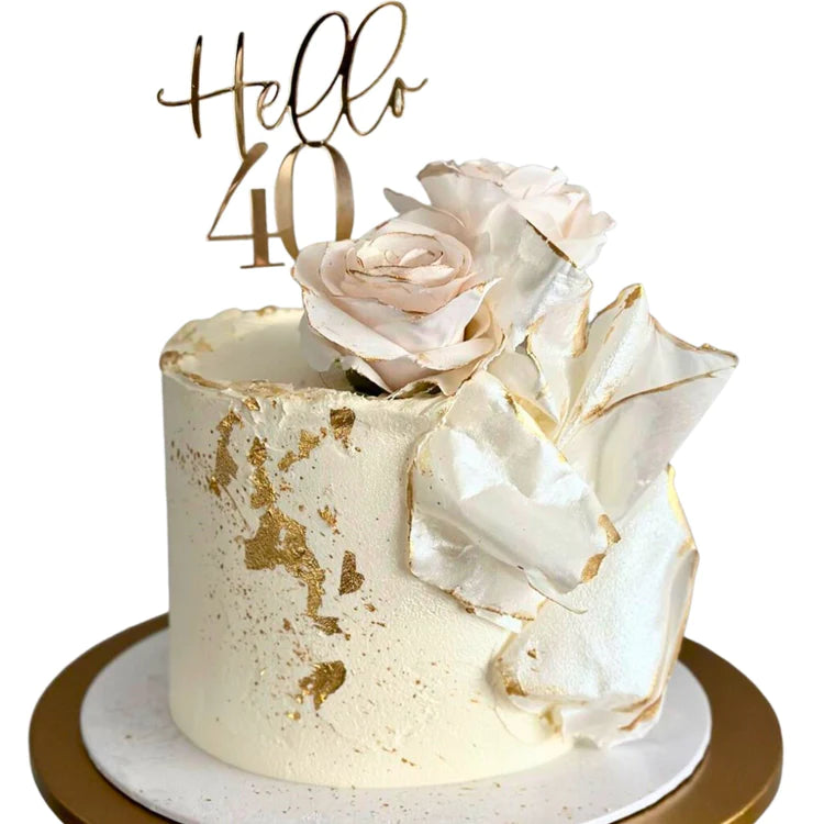 A decorative cake featuring the words "Hello 40" in a gold acrylic topper. The cake is cream-colored with a textured surface and has gold leaf decoration. It is adorned with faux pink roses and a shiny, sculpted white element. The cake sits on a circular white and gold cake stand.