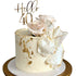 A decorative cake featuring the words "Hello 40" in a gold acrylic topper. The cake is cream-colored with a textured surface and has gold leaf decoration. It is adorned with faux pink roses and a shiny, sculpted white element. The cake sits on a circular white and gold cake stand.