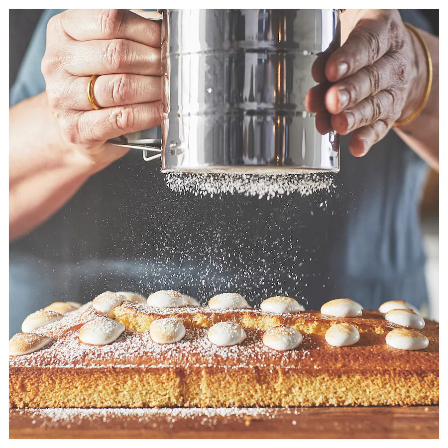 A person is holding a stainless steel flour sifter above a baked dessert, sprinkling powdered sugar onto the surface. The sifter is cylindrical with a handle, and fine sugar particles are falling onto a rectangular cake topped with small round decorations. The background features a blurred kitchen setting, adding a warm ambiance to the scene.